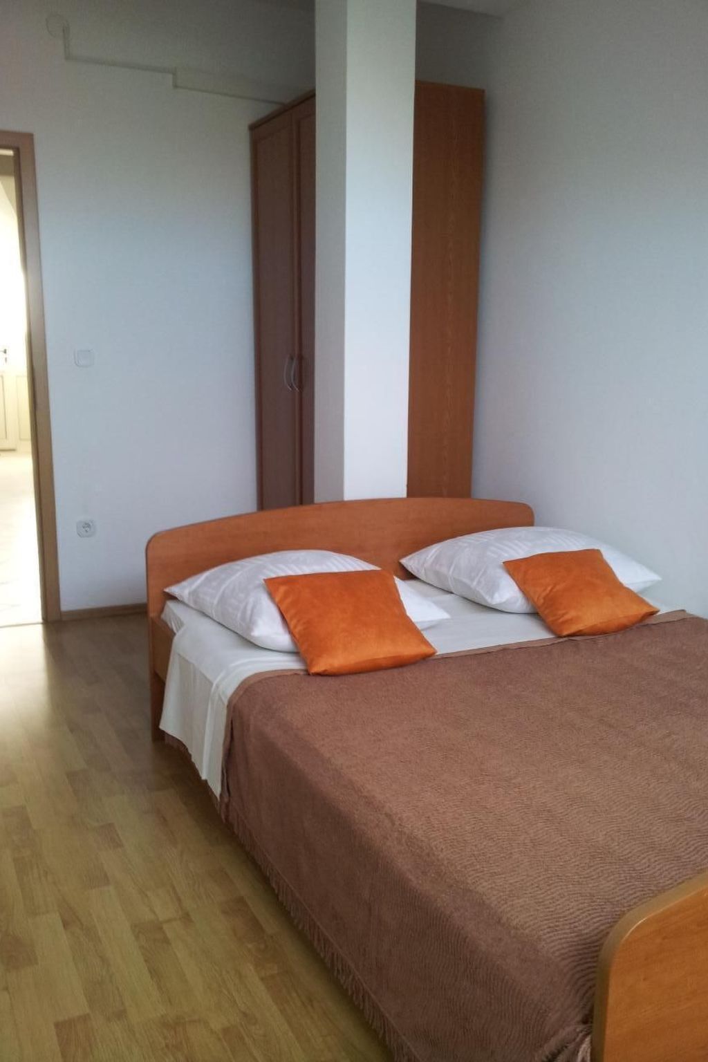 Een slaapkamer met een bed, witte en oranje kussens, een bruine plaid en een houten garderobe.