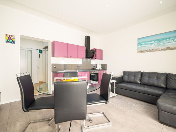 A modern living room with a black leather sofa, a glass dining table with chairs, a kitchen with pink cabinets, and a painting of a beach scene. - Apartment Sukosan by Interhome (Debeljak)