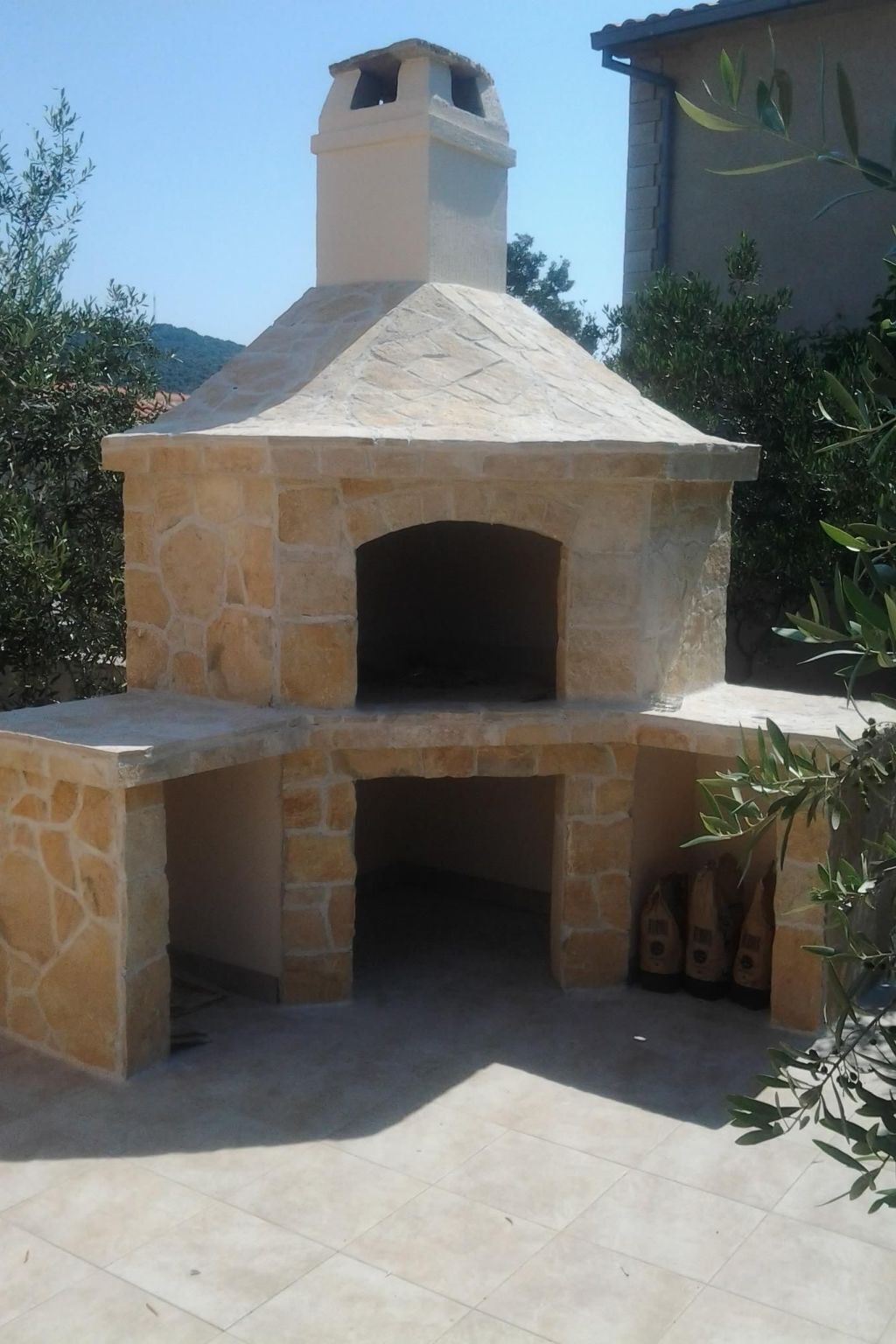 A stone-built outdoor pizza oven with a chimney, surrounded by a tiled patio and greenery.