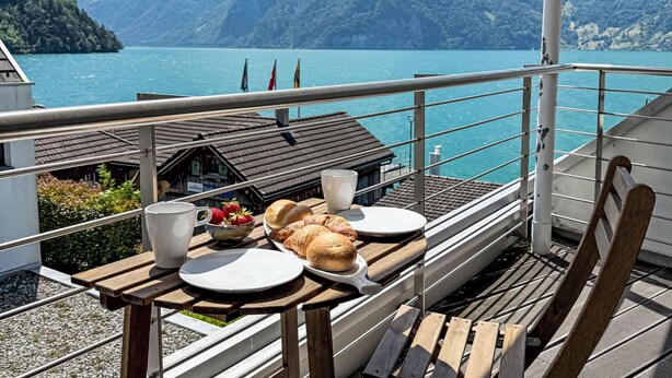 A balcony with a table set for breakfast, overlooking a lake and mountains.