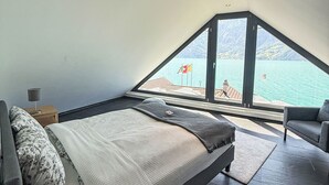 A bedroom with a bed, a chair, a nightstand, and a view of a lake and mountains through a large window.