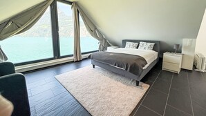 A bedroom with a bed, a nightstand, a lamp, a chair, and a view of the water. - Villa am See 5 - Beach House by Interhome (Sisikon)