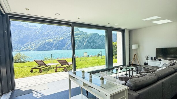 A modern living room with a large glass door leading to a balcony overlooking a lake and mountains. - Villa am See 5 - Beach House by Interhome (Sisikon)