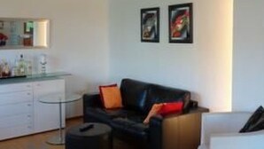 Una sala de estar moderna con un sofá de cuero negro, un sillón blanco y una mesa de centro de vidrio. Se observan dos obras de arte enmarcadas en la pared y una zona de cocina con armarios blancos y una encimera.