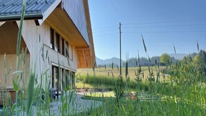 A modern wooden house with large windows and a sloped roof, surrounded by tall grass and a clear sky.