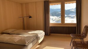 A bedroom with a bed, a desk lamp, a chair, and a window with a view of mountains.