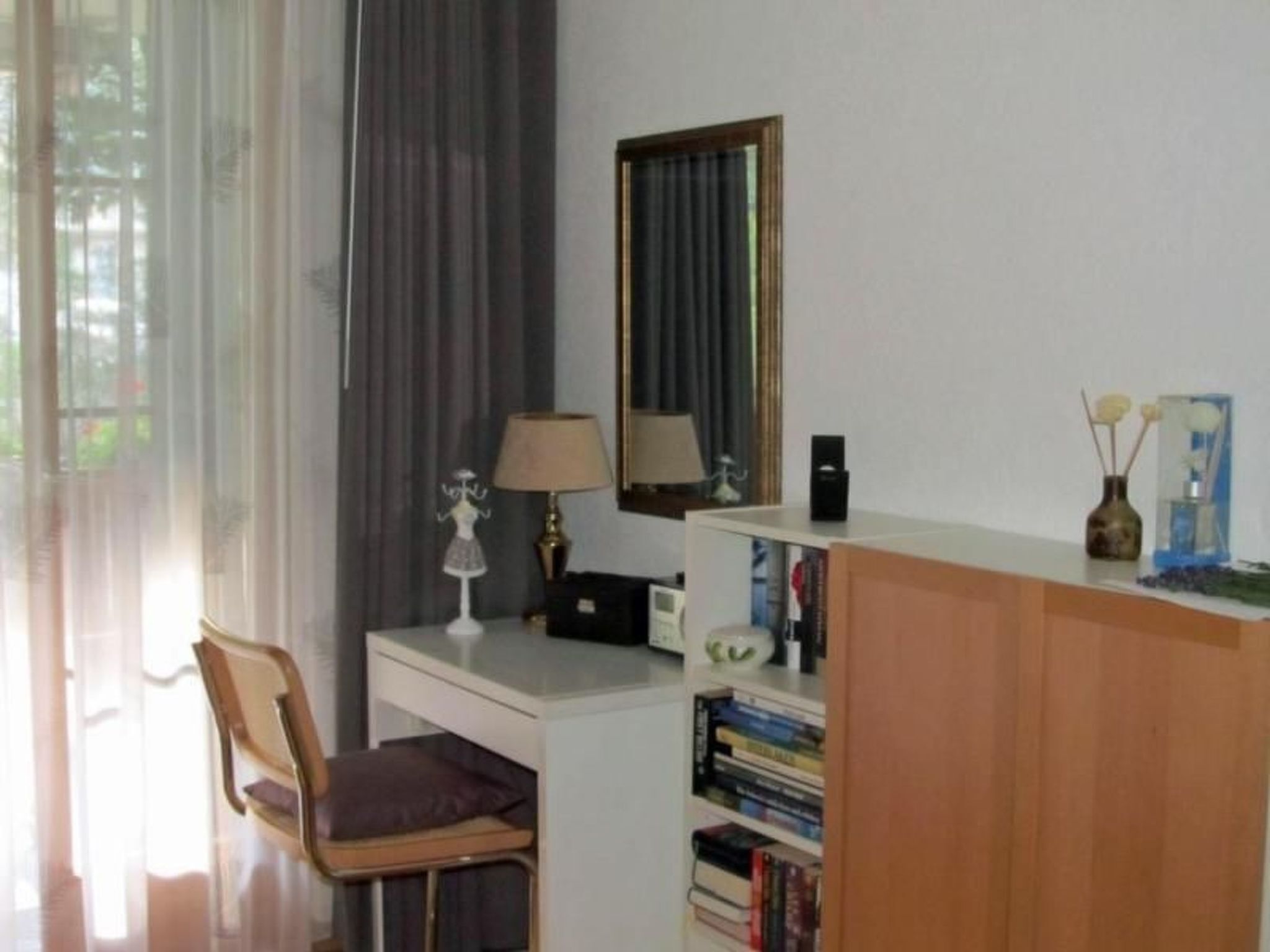 A room with a desk, chair, books, a lamp, a mirror, and a window with curtains.