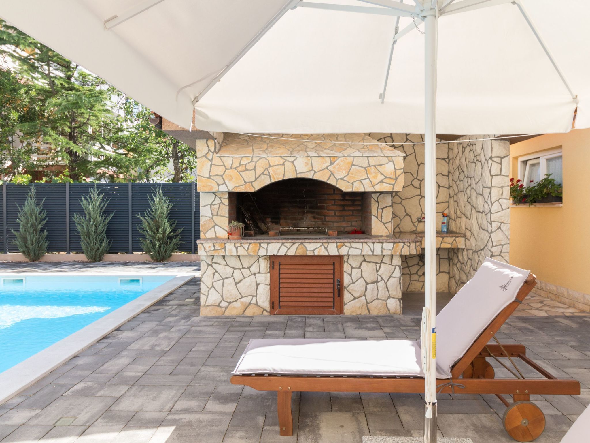 Een buitenkeuken van natuursteen met een barbecue, een zwembad en een lounge stoel onder een overkapping.