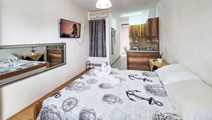 Un dormitorio con cama, espejo, televisión, una mesita pequeña y una kitchenette al fondo.