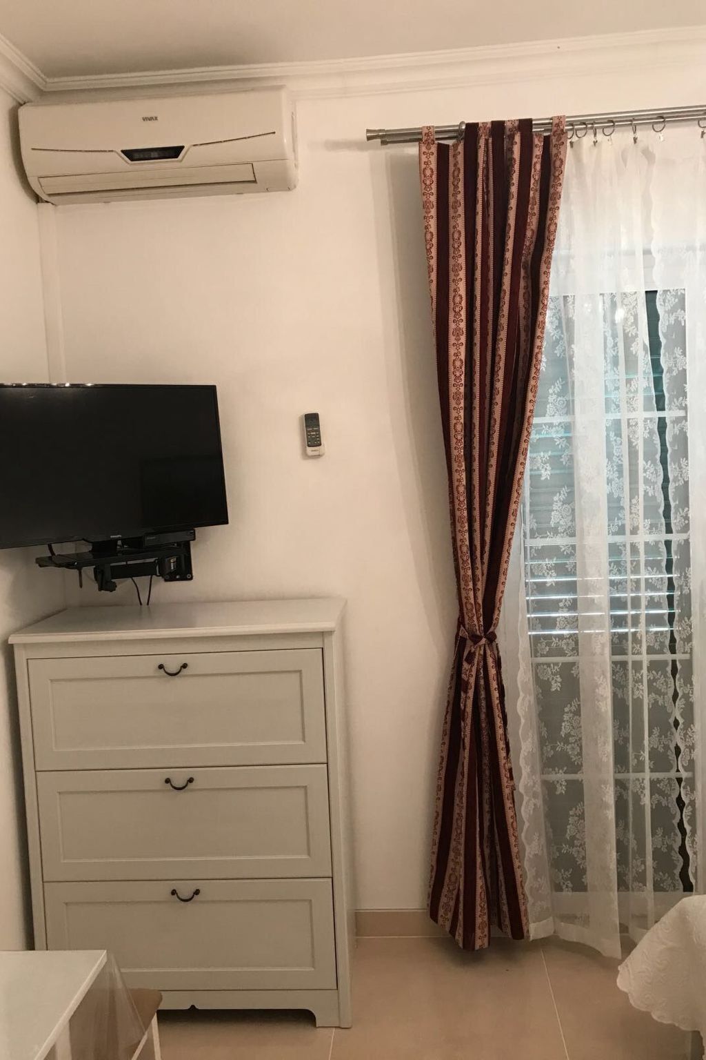 Habitació amb televisió de pantalla plana penjada en una petita taula blanca, aire condicionat fixat a la paret i una finestra amb cortines estampades.