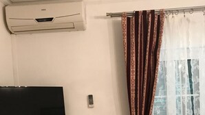 Ein Zimmer mit einem an einer weißen Kommode befestigten Flachbildfernseher, einer Wandklimaanlage und einem Fenster mit gemusterten Vorhängen.