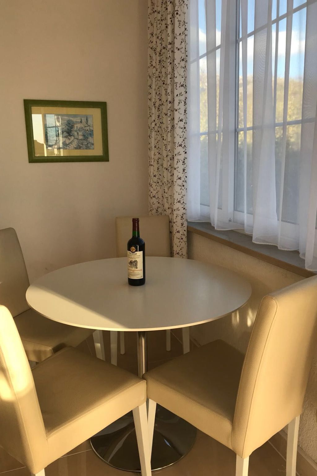 Un coin repas avec une table ronde et quatre chaises de couleur beige, une bouteille de vin, un tableau encadré au mur et une fenêtre ornée de rideaux légers.