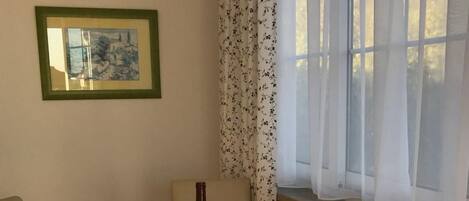 Un coin repas avec une table ronde et quatre chaises de couleur beige, une bouteille de vin, un tableau encadré au mur et une fenêtre ornée de rideaux légers.