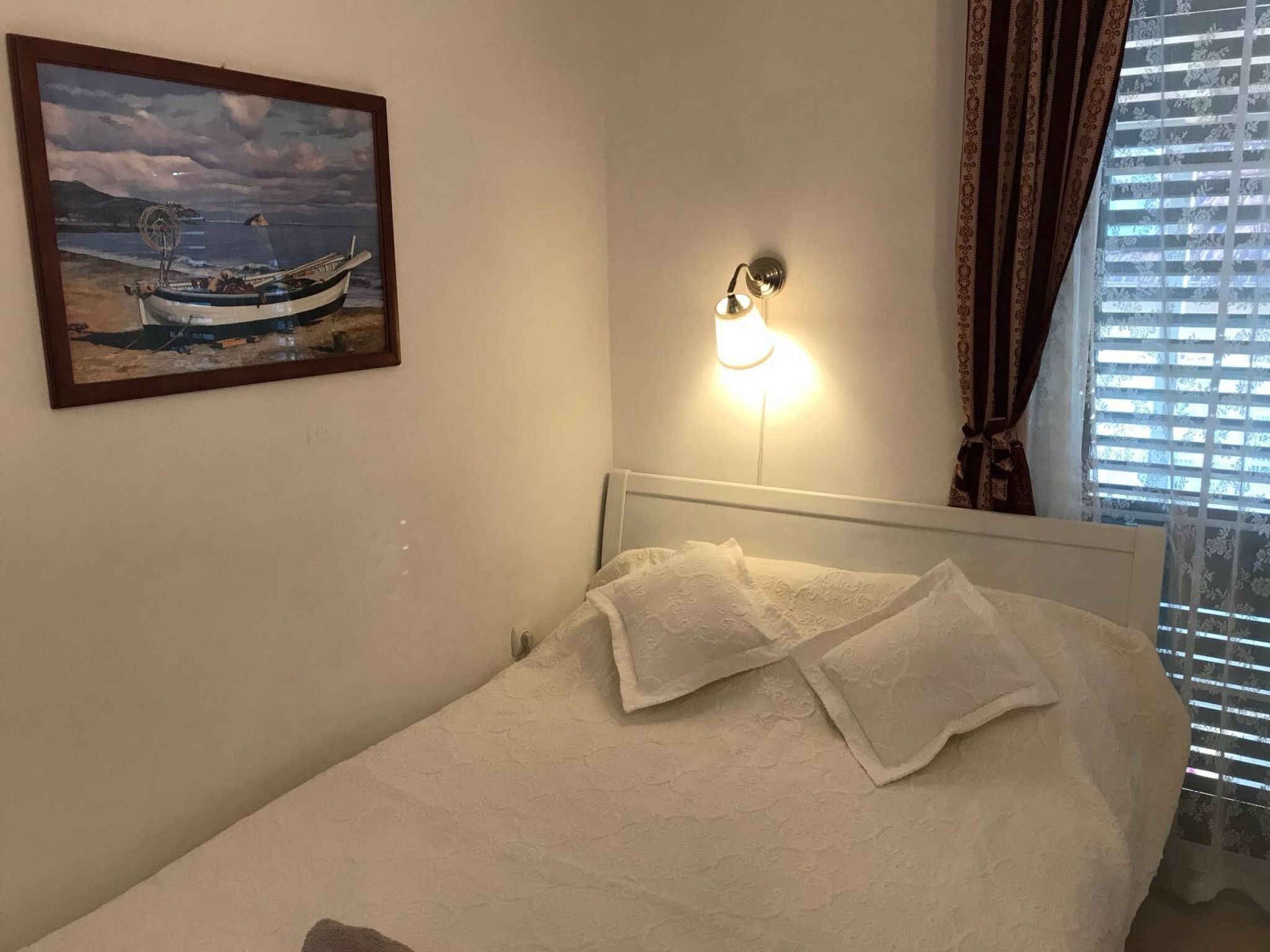 A bedroom with a bed, a wall-mounted light, a framed painting of a boat, and a window with blinds.