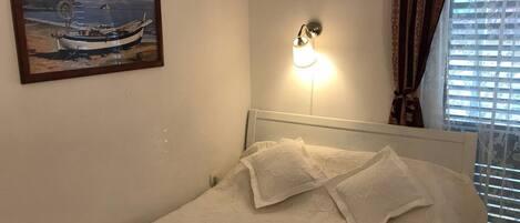 A bedroom with a bed, a wall-mounted light, a framed painting of a boat, and a window with blinds.