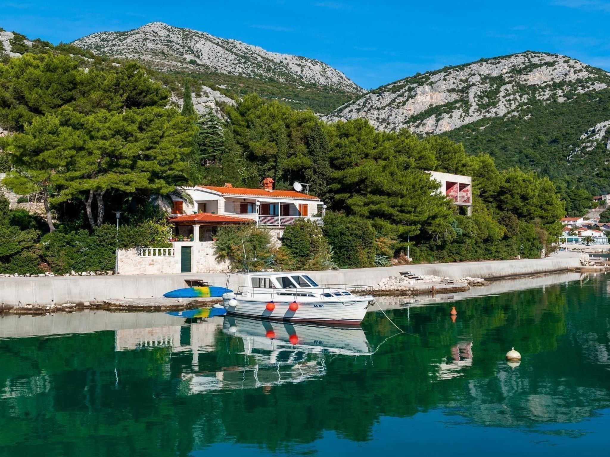 A coastal building with a balcony, a boat docked nearby, and mountains in the background.