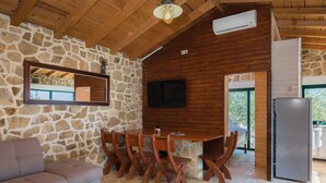 Ein gemütliches Zimmer mit Steinwand, Holzboden, einem an der Wand montierten Fernseher, einem Esstisch mit Stühlen, einer Couch und einem Kühlschrank.