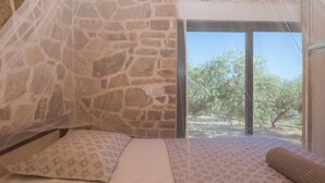 Ein Zimmer mit Steinsäulen, einem Bett, einem Moskitonetz und Blick auf Bäume.