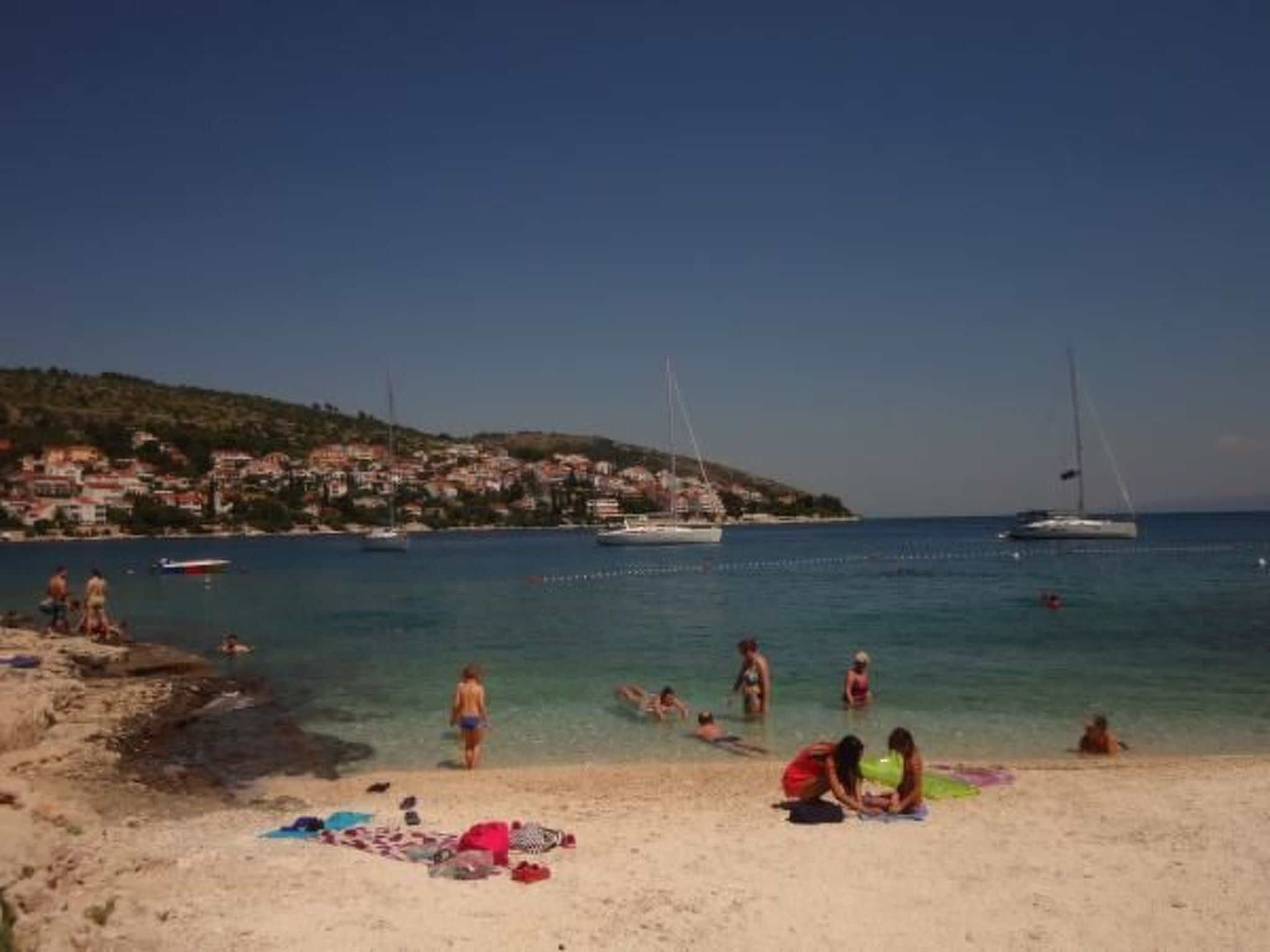 A beach with people sunbathing and swimming, boats in the water, and a hillside with buildings in the background.