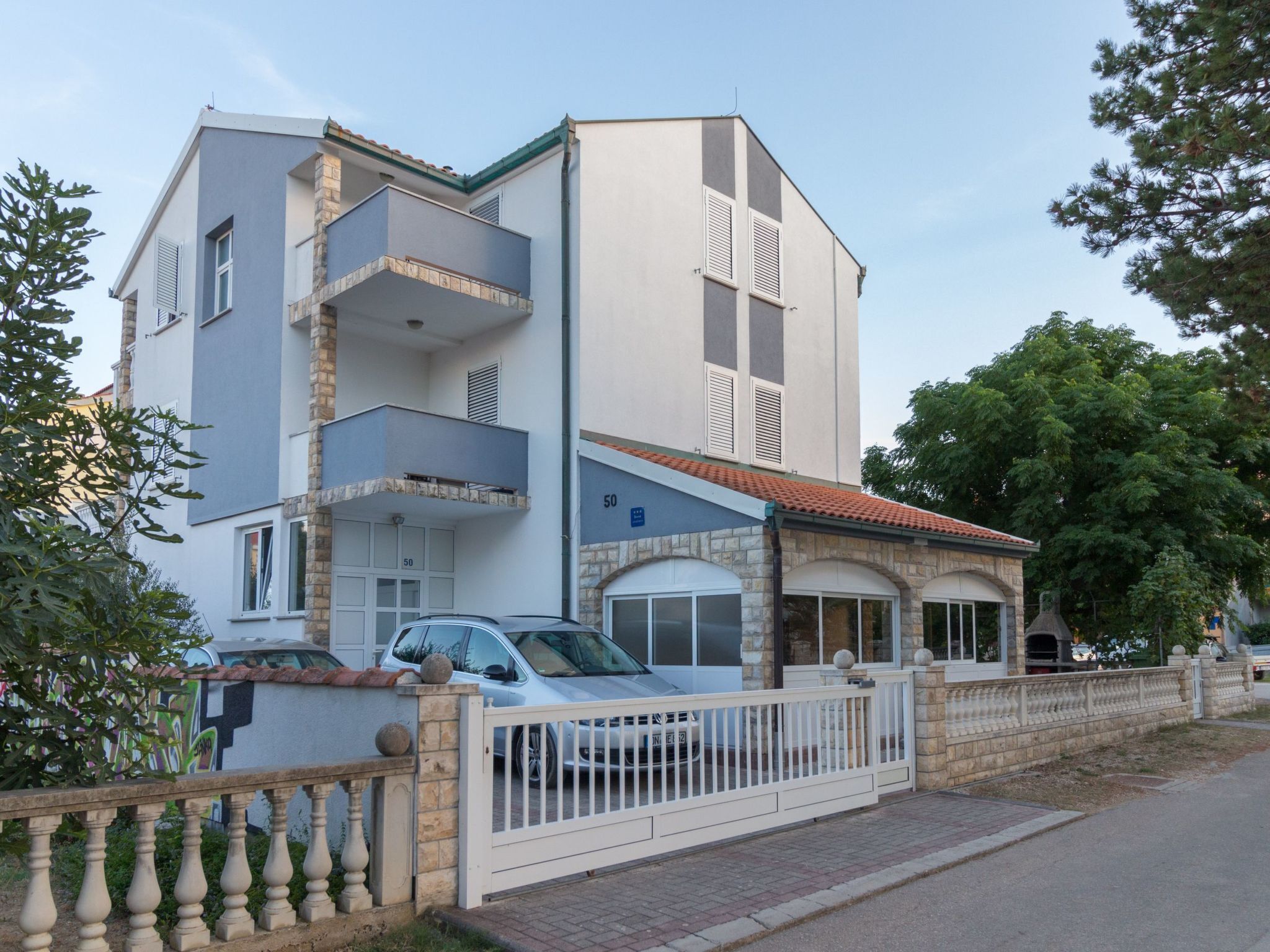 Ein zweistöckiges Gebäude mit einer Steinfassade und einem Balkon im oberen Geschoss, sowie eine Einfahrt mit Tor.