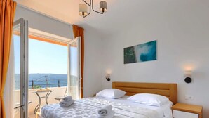 Ein Hotelzimmer mit Bett, Nachttisch, einem Gemälde und Blick auf das Meer durch eine offene Tür.