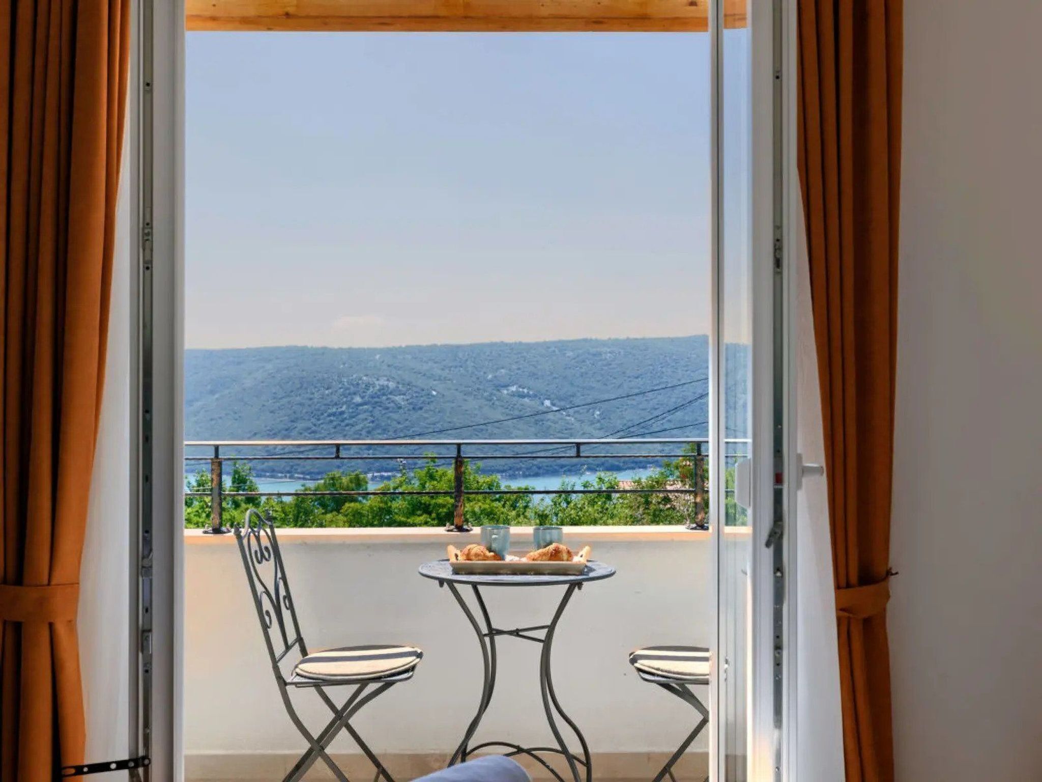 Ein Balkon mit zwei Stühlen und einem kleinen Tisch, mit Blick auf eine schöne Landschaft aus Hügeln und einem Gewässer.