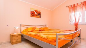 Una cama individual con colcha naranja en una habitación con paredes color melocotón, una mesita de noche con lámpara y una ventana con cortinas translúcidas.