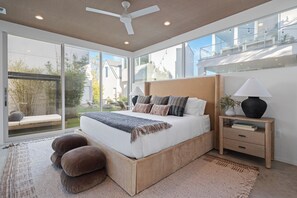 Ein modernes Schlafzimmer mit einem großen Bett, einem Nachttisch, einem Deckenventilator und Blick ins Freie.