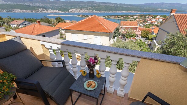 A balcony with two black chairs, a small table with a plate of food, and a vase with flowers. A scenic view of a coastal town with red-roofed buildings and a body of water is visible in the background. - Pension Tina Appartement 2 by Interhome (Krk/Šilo)