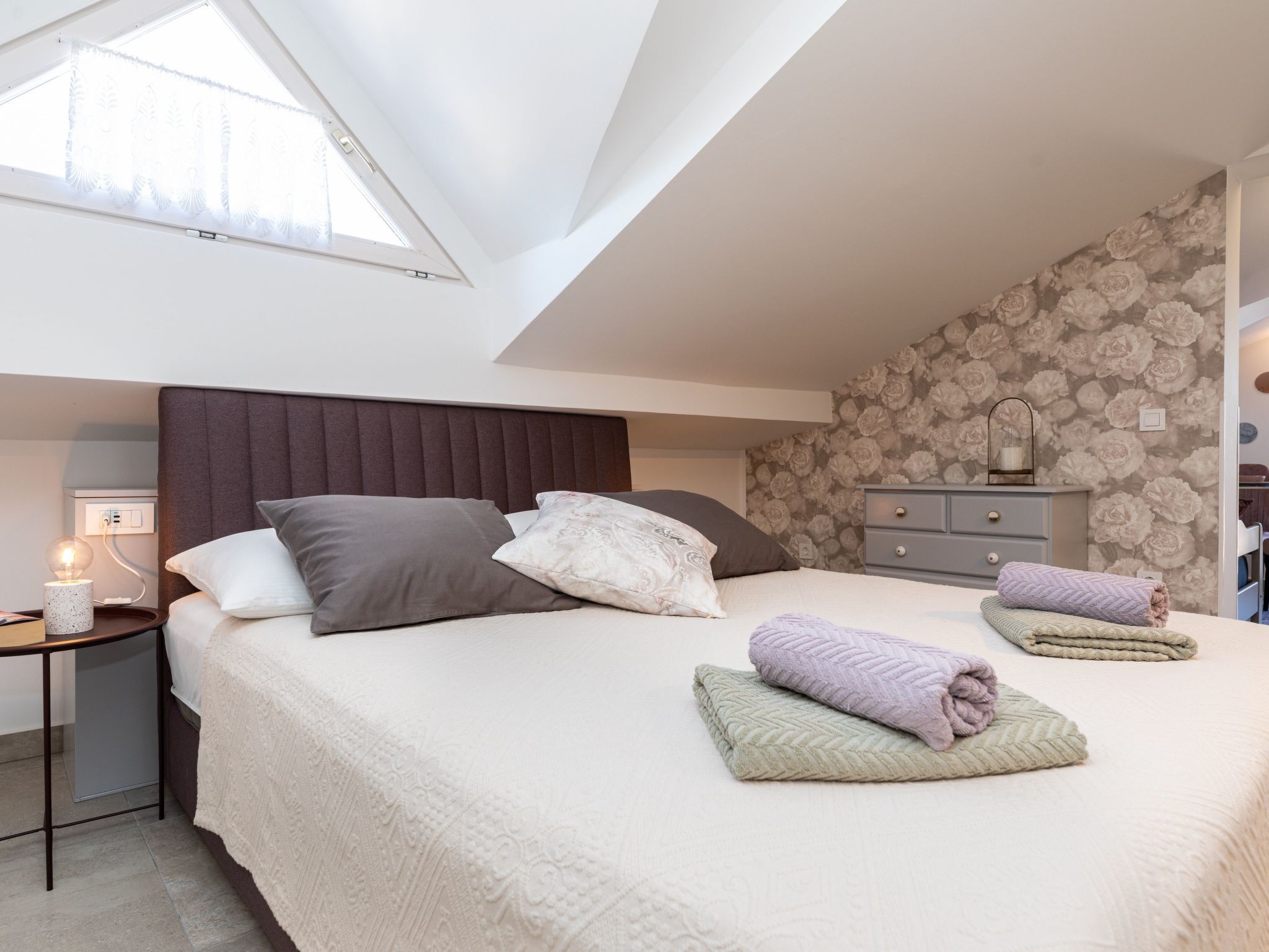Ein Schlafzimmer mit schrägverdeckter Decke, einem Bett mit Kopfteil, einem Nachttisch mit Lampe, einer Kommode und einer Wand mit Blumenmuster-Tapete.