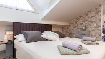Ein Schlafzimmer mit schrägverdeckter Decke, einem Bett mit Kopfteil, einem Nachttisch mit Lampe, einer Kommode und einer Wand mit Blumenmuster-Tapete.