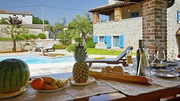 A poolside table with fruits, a watermelon, and a pineapple, overlooking a pool and a stone building with blue shutters. - Vacation home Simone by Interhome (Dracevac)
