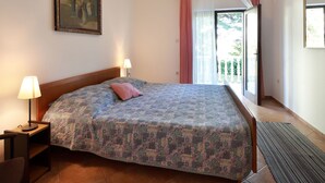 A bedroom with a large bed, a nightstand with a lamp, a painting on the wall, and a window with curtains. - Svoboda 2 by Interhome (Krk/Šilo)