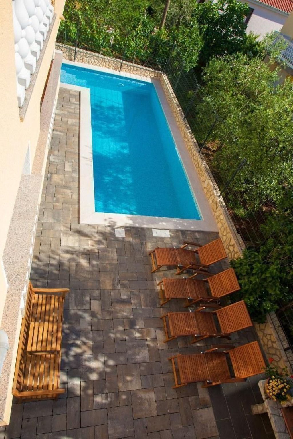 A rectangular swimming pool with a stone border, surrounded by a stone patio and wooden lounge chairs.
