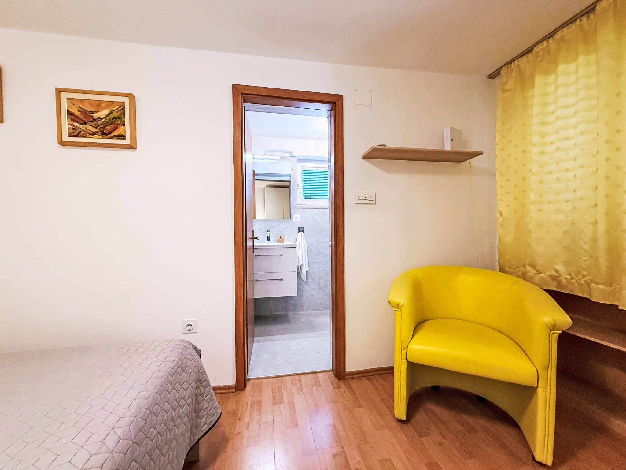 A room with a bed, a yellow chair, a wooden door leading to a bathroom, and a small shelf.
