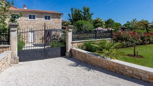 Una casa de piedra con entrada protegida por una reja, camino de grava y jardín cuidado con diversas plantas y una palmera.