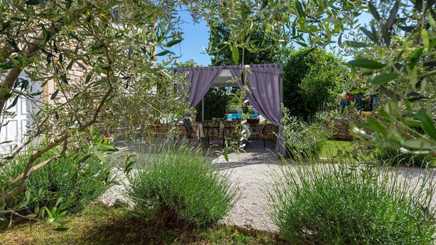 Un espacio al aire libre con pérgola, mesas y sillas, rodeado de follaje y un sendero de grava.