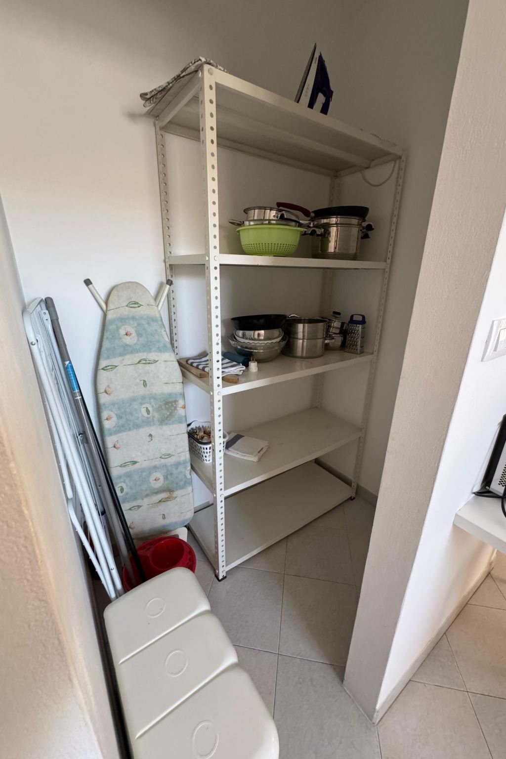 Une cuisine avec une étagère en métal où sont rangés des casseroles, des poêles et une passoire. On y aperçoit un appareil blanc, qui pourrait être un grille-pain, et une planche à repasser rayée appuyée contre le mur.