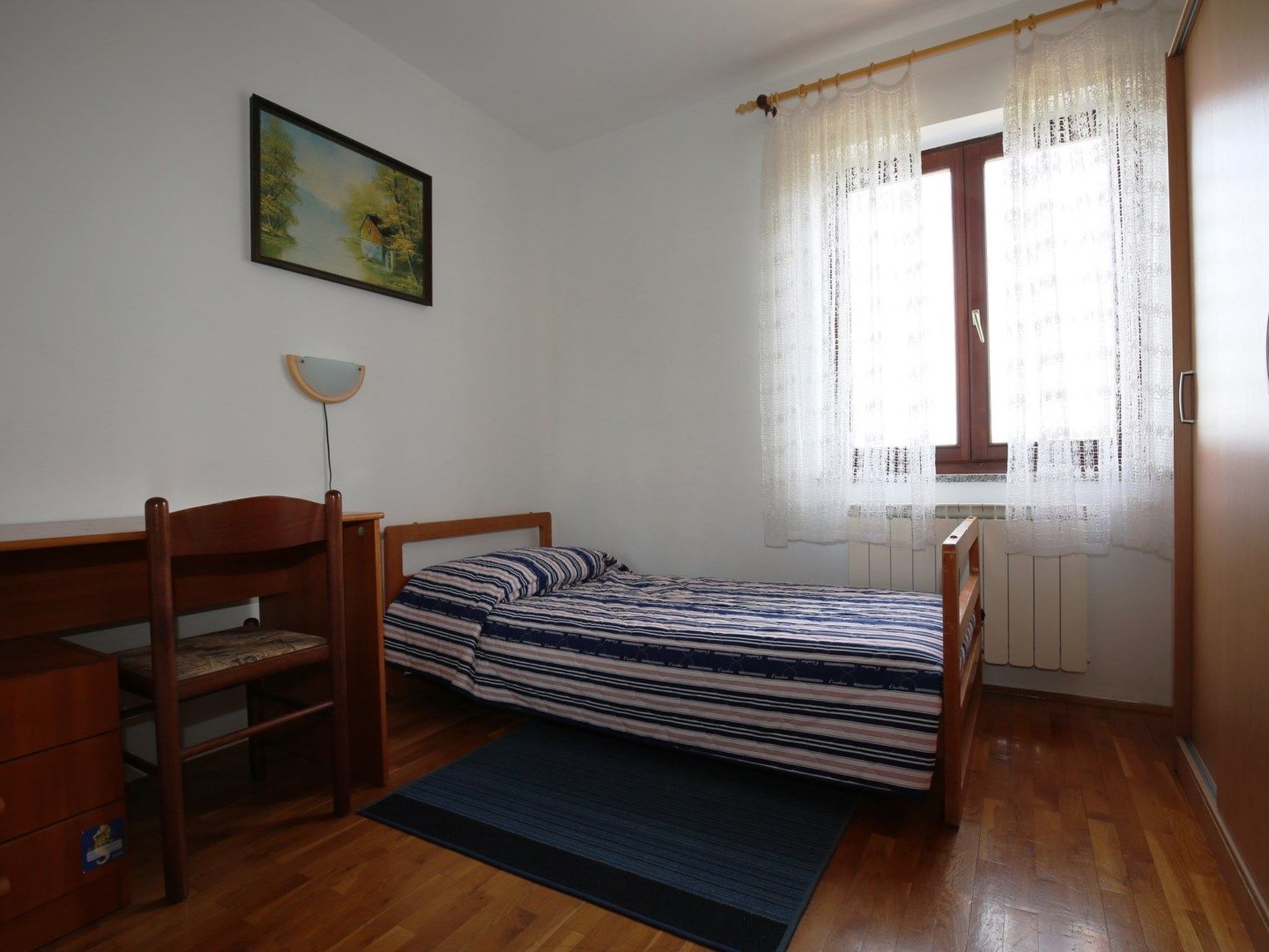 Ein Schlafzimmer mit einem Einzelbett, einem Holztisch, einem Stuhl, einem Heizkörper und einem Fenster mit durchsichtigen Vorhängen.