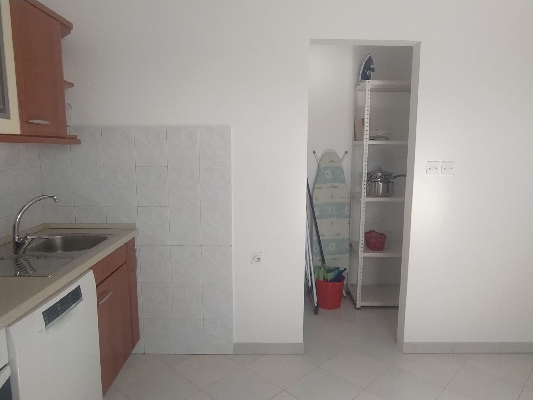 A kitchen with a dishwasher, a sink, and a cabinet. There is a red bucket on the floor and a shelf with a pot and a red container.