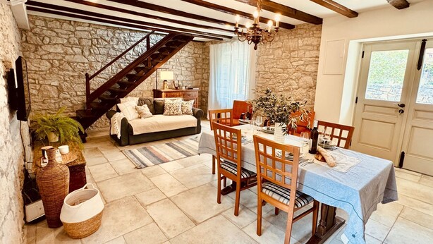 Una habitación acogedora con pared de piedra, vigas de madera, una escalera, una mesa de comedor con sillas y un candelabro.