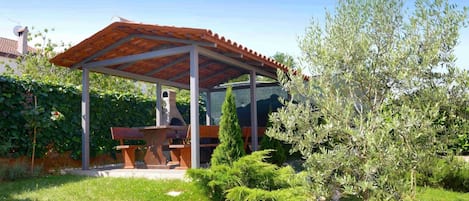 Uma área de estar coberta ao ar livre, com mesa e bancos, cercada por vegetação e uma árvore.