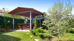 Un'area salotto all'aperto riparata, con un tavolo e panche, immersa nel verde e con un albero.
