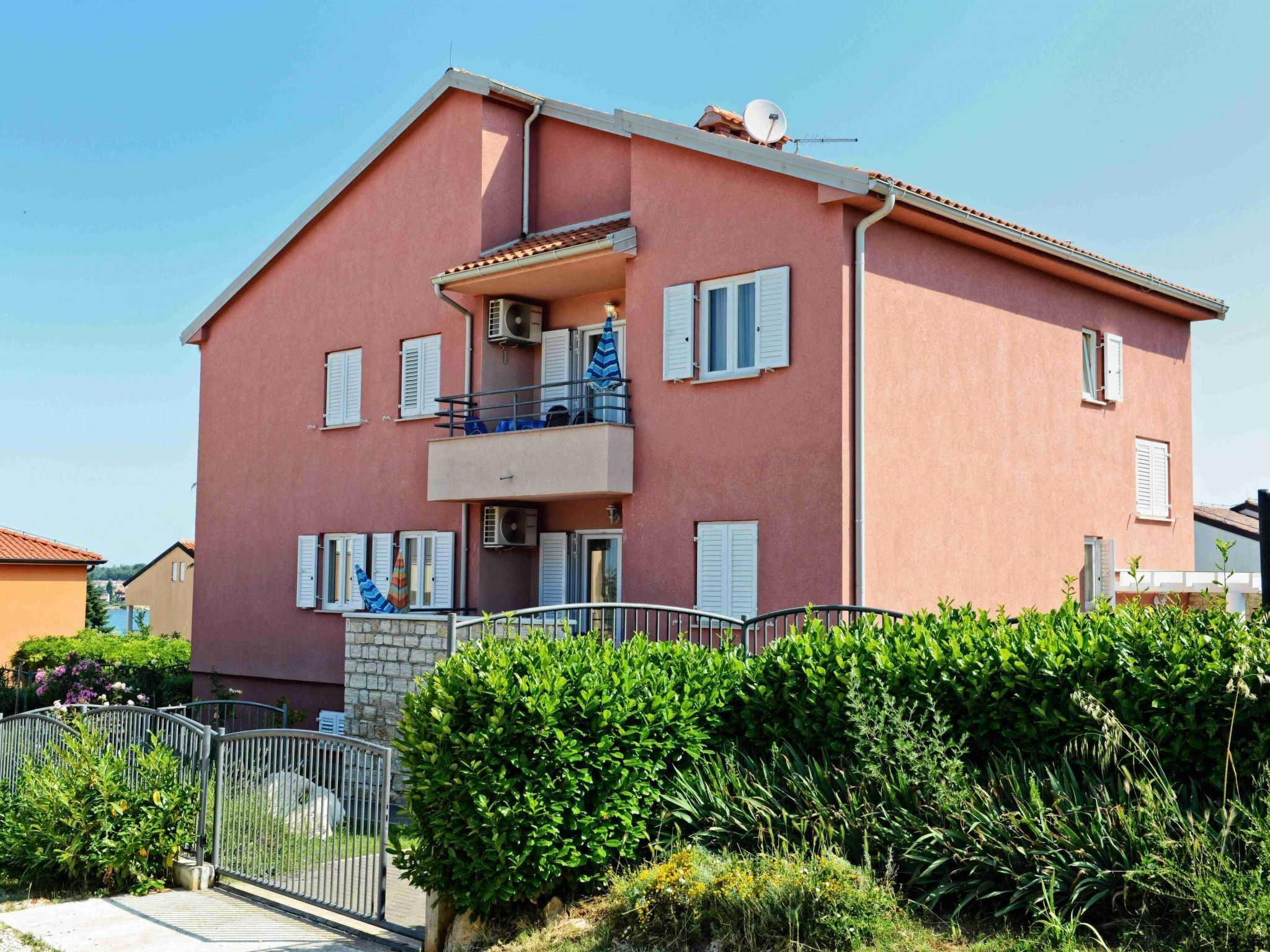 Un edificio su due piani con un balcone, un ingresso chiuso da cancello e vegetazione circostante.