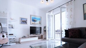 Un soggiorno moderno con un tavolino in vetro, un divano grigio, una TV a schermo piatto su un mobile bianco, una chitarra e una libreria con libri e oggetti decorativi.