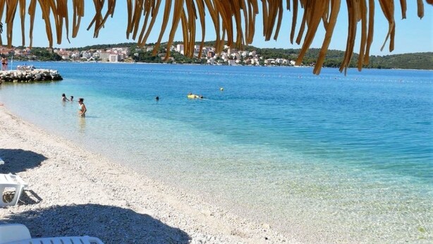 Una spiaggia con acque cristalline, una struttura con tetto di paglia e una costa lontana con edifici.