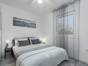 Een slaapkamer met een bed, nachtkastje, lamp, een foto aan de muur en een raam met gordijn.