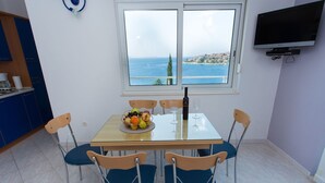 Uma sala de jantar com uma mesa posta para dois, uma cesta de frutas, duas taças de vinho e uma garrafa de vinho. Há uma janela com vista para o mar e uma televisão fixada na parede.