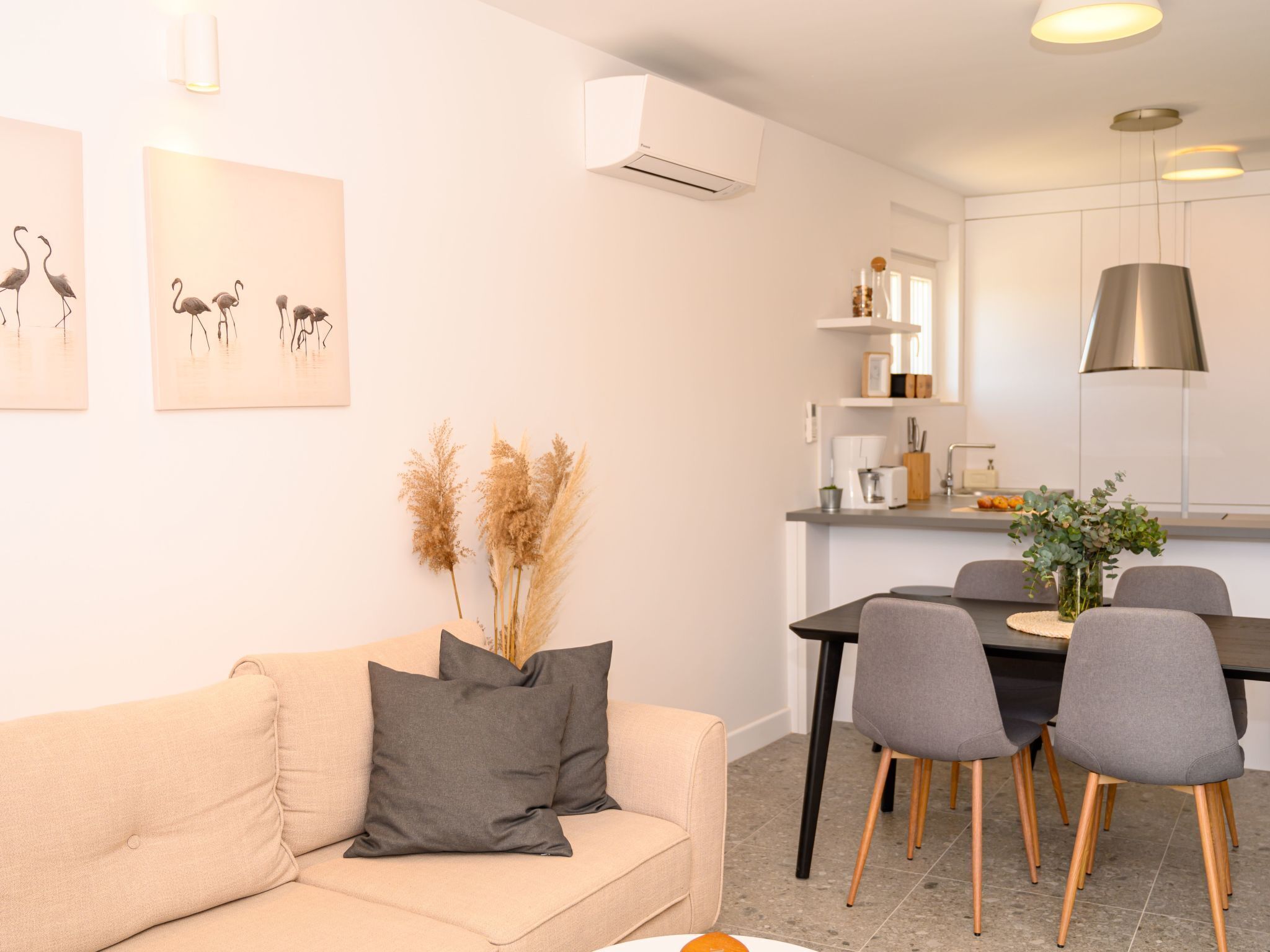 Un salon moderne avec un canapé beige, une table à manger avec des chaises et un coin cuisine doté de meubles blancs et d’une hotte de cuisine en acier inoxydable.
