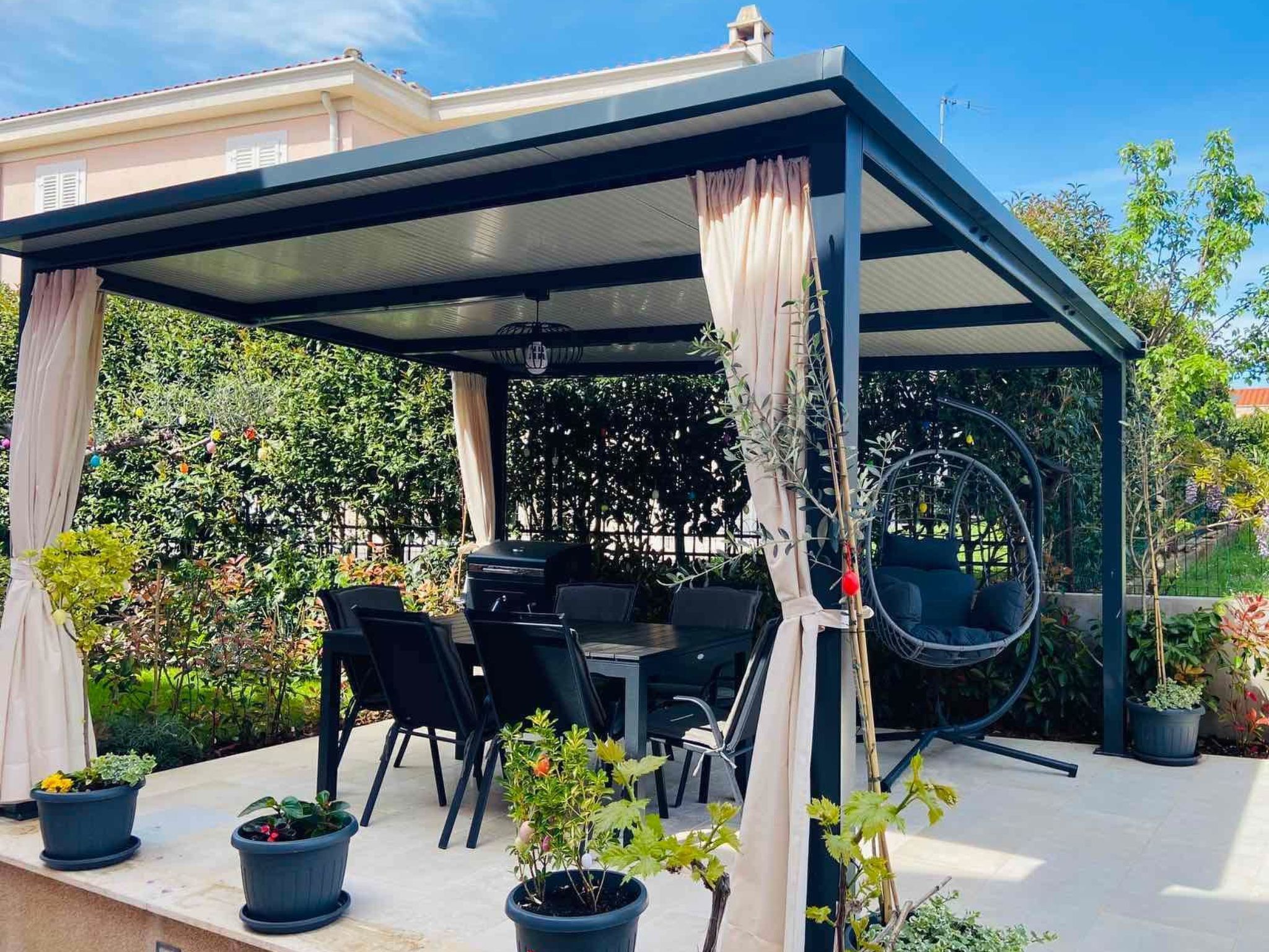 Een overdekt terras met een tafel en stoelen, omgeven door potplanten en een hangstoel.